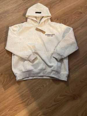 Grå Essentials Fear of God hoodie - Snygg ljusgrå hoodie från Essentials Fear of God med svart logga tryckt på bröstet. Klassisk passform med huva och ribbade muddar vid ärmar och nederkant. Perfekt för en avslappnad och trendig streetwear-look.