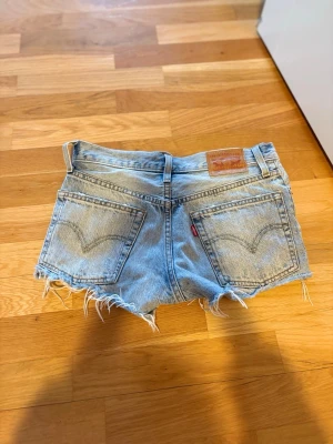 Levi's ljusblå jeansshorts med fransar - Säljer ett par klassiska ljusblå jeansshorts från Levi's med råa, fransiga kanter och slitna detaljer. Shortsen har 4 fickor, knappgylf och Levi's ikoniska läderpatch bak. Perfekta för sommaren och riktigt snygga till sneakers eller sandaler.