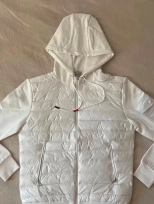 Vit pufferjacka med huva från Moncler - Säljer en stilren vit pufferjacka från Moncler med huva och dragkedja framtill. Jackan har två fickor med dragkedja och ribbade muddar vid ärmsluten. Materialet är quiltat och ger en modern look. Perfekt för kalla dagar och snygg till alla outfits.