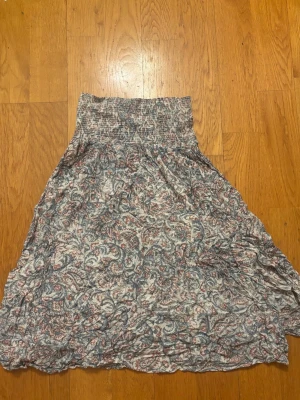 Mönstrad långkjol med smockmidja - Luftig långkjol med smockad midja och ett detaljerat blommigt mönster i blått, rött och vitt. Kjolen har en avslappnad passform och är tillverkad i ett mjukt, tunt material som passar perfekt till varmare dagar.