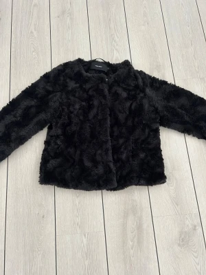 Svart fuskpälsjacka från Vero Moda - Mjuk och fluffig svart fuskpälsjacka från Vero Moda med rund halsringning och tryckknappar framtill. Jackan har helfodrad insida och en rak, avslappnad passform som ger en trendig look. Perfekt för kalla dagar när du vill vara både varm och snygg. Storlek XL men passar mig som har S. 