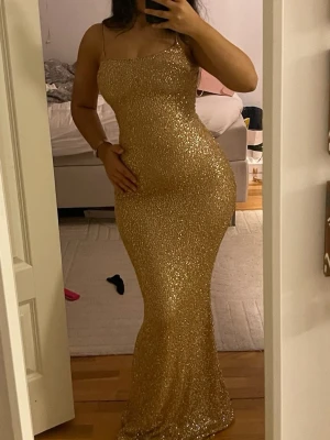 Glittrig guldig långklänning - Säljer en superglittrig guldig långklänning från ohpolly med smala axelband och figurnära passform. Klänningen har en snygg, kroppsnära siluett och är täckt av glittrande paljetter som verkligen fångar ljuset. Perfekt för att sticka ut på bal eller fest! Klänningen är i väldigt bra skick och har endast varit använd i ett par timmar.