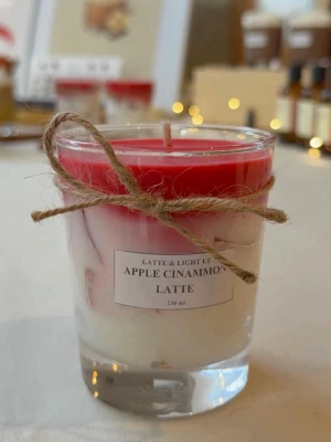 Apple Cinnamon Latte doftljus - Säljer ett handgjort doftljus i glas med etiketten 'Apple Cinnamon Latte'. Ljuset har en snygg röd och vit swirl-design och är dekorerat med ett snöre runt glaset. Perfekt för att skapa mysig stämning hemma. Volym: 230 ml.