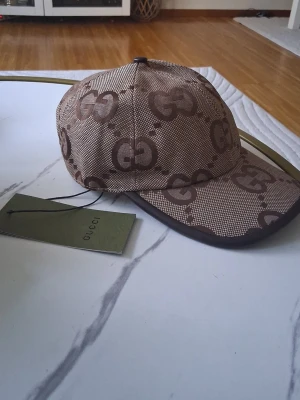 Brun keps med GG-mönster från Gucci - Säljer en brun keps från Gucci med det klassiska GG-monogrammet i mörkbrunt över hela kepsen. Kepsen har böjd skärm och är tillverkad i ett slitstarkt tyg med läderdetaljer runt skärmen. Perfekt för dig som vill ha en exklusiv och trendig accessoar. Helt ny skick!!!