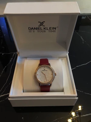 Daniel Klein klocka med rött band - Stilren klocka från Daniel Klein med ett vinrött band och guldfärgat boett. Urtavlan är vit med subtilt mönster och kantad av gnistrande stenar. Perfekt accessoar för dig som vill ha något elegant och trendigt. Boxen följer med och ett helt nytt batteri utöver det som är i klockan. Nypris runt 900kr. Priset kan diskuteras! 