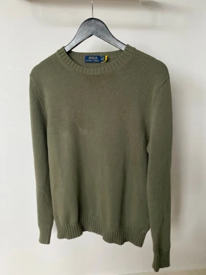 Olivgrön stickad tröja Polo Ralph Lauren - Snygg olivgrön stickad tröja från Polo Ralph Lauren med rund halsringning och diskret logga på bröstet. Tröjan har ribbade muddar vid ärmslut och nederkant. Perfekt för lager på lager under kyliga dagar.