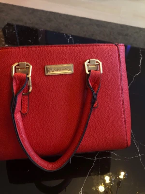 Lyxig röd handväska  - Snygg röd handväska med guldfärgade metalldetaljer och dubbla handtag. Väskan är tillverkad i strukturerat syntetmaterial och har en dragkedja på baksidan. Perfekt för dig som vill sticka ut med en färgstark accessoar. 