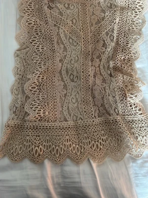 Beige spetsblus med mönster - Supersnygg beige blus i spets med detaljerat blommigt och geometriskt mönster. Blusen har vågiga kanter och transparent material som ger en romantisk och trendig look. Perfekt att styla lager på lager. Materialet känns lätt och luftigt.