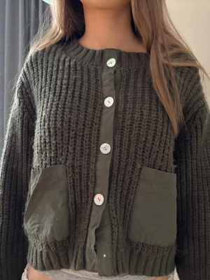 Zara Olivgrön stickad kofta med fickor - Säljer en olivgrön stickad kofta med stora framfickor i tyg och vita knappar. Koftan har grovstickad struktur, rund halsringning och ribbade muddar. Tyvärr saknar den 1 knapp längst ned 
