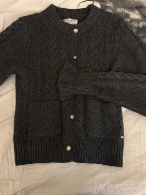 Mörkgrå kabelstickad kofta  - Mysig mörkgrå kabelstickad kofta med söta detaljer, två framfickor och söta ärmar. Koftan har ribbade muddar och nederkant samt ett klassiskt flätmönster. Nästan aldrig använd så i nyskick, köptes för ca 500kr