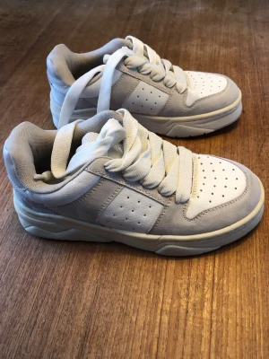 Vita och grå chunky sneakers - Snygga chunky sneakers i vitt och ljusgrått med breda vita skosnören och perforerade detaljer på tån och sidorna. Ovandelen är i en mix av mocka och syntetmaterial. Knappt använda.
