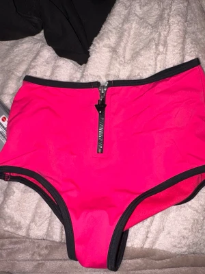 Neonrosa bikiniunderdel från Guess - Säljer en snygg neonrosa bikiniunderdel från Guess med hög midja och svart dragkedja framtill. Bikinin har svarta kanter och Guess-logga baktill. Materialet är stretchigt och snabbtorkande, perfekt för stranden eller poolen.