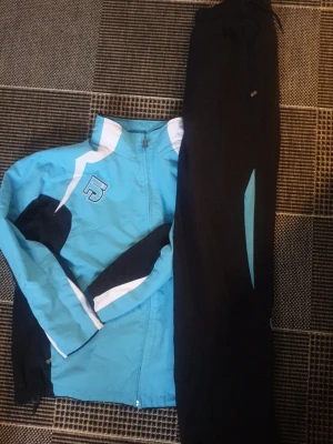 Blått träningsset Five jacket & byxor - Säljer ett sportigt träningsset från Five med blå och svarta detaljer. Jackan har dragkedja, vit logga på bröstet och vita inslag på ärmarna. Byxorna är svarta med blå detalj längs benet. Perfekt för löpning eller träning utomhus.