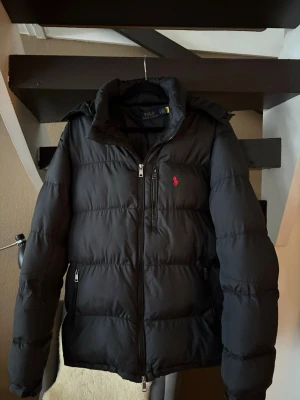 Svart pufferjacka från Polo Ralph Lauren - Snygg svart pufferjacka från Polo Ralph Lauren med klassisk röd logga på bröstet. Jackan har hög krage, två sidofickor med dragkedja och en rak passform. Perfekt för kalla vinterdagar och riktigt stilren look. Storlek S men passar även M 