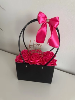 Svart dekorväska med rosa rosetter - En svart dekorativ väska med rundade handtag och en stor rosa rosett upptill. Väskan är fylld med konstgjorda rosa rosor och har en 'Happy Birthday'-skylt i guld. Perfekt som present eller dekoration.