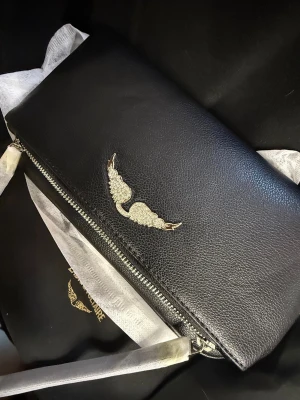 Svart handväska Zadig & Voltaire - Snygg svart handväska från Zadig & Voltaire i äkta läder. Väskan har ett elegant silverfärgat vinge-emblem på framsidan och dragkedja upptill. Stilren och enkel design med justerbar axelrem. Perfekt för dig som gillar minimalistisk och trendig look.