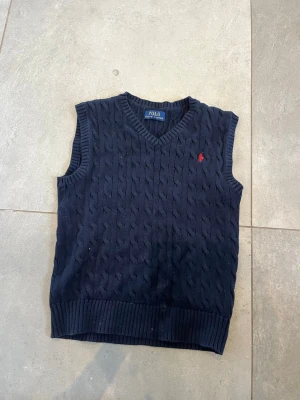 Mörkblå kabelstickad väst Polo Ralph Lauren - Kabelstickad mörkblå väst från Polo Ralph Lauren med V-ringning och ribbade kanter. Klassisk röd broderad logga på bröstet. Perfekt för lager på lager och ger en preppy look. Tillverkad i mjukt stickat material. Helt ny kostar 2100 från boozt och den är i bra skick använt max 6-7 gånger