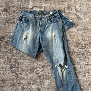 Blå low rise straight jeans med slitningar - Säljer ett par blå jeans med low rise och straight fit. Byxorna har tydliga slitningar och hål på både fram- och baksida, vilket ger en cool och avslappnad vibe. 39 cm i midjan tvärs över och 75 cm innerben