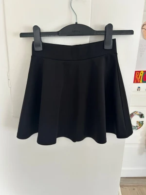 Svart kortkjol i A-linje - Säljer en svart kortkjol i A-linjeform med bred linning. Kjolen har en enkel och stilren design som passar till det mesta. Materialet känns mjukt och har ett lätt stretchigt fall. Från Kappahl i storlek 134/140, vilket är typ XS ❤️