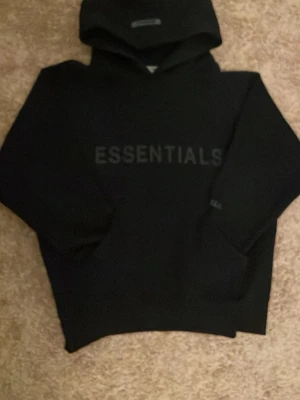 Svart Essentials Fear of God hoodie - Snygg svart hoodie från Fear of God Essentials med stor logga på bröstet och mindre tryck på ärmen. Klassisk passform med huva och ribbade muddar. Tillverkad i mjukt bomullsmaterial som känns riktigt skönt mot huden. Kom gärna med bud!!