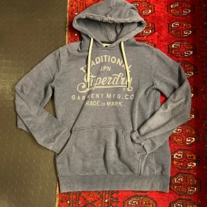 Grå hoodie från Superdry   - Säljer en grå hoodie från Superdry med stort vintage-inspirerat tryck på bröstet. Tröjan har huva med vita snören och en klassisk känguruficka framtill. Mjuk insida och ribbade muddar vid ärmslut och nederkant. Perfekt för chill dagar. Står inte storleken i men runt xs/s nånstans där 