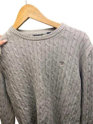Grå kabelstickad tröja från Gant - Kabelstickad grå tröja från Gant med rund halsringning och diskret broderad logga på bröstet. Tröjan har klassisk passform och är tillverkad i mjukt material som passar perfekt till lager-på-lager-stil. 