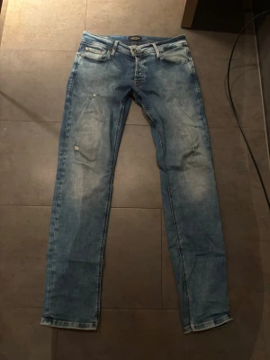 Slim fit jeans från Jack & Jones - Snygga ljusblå slim fit jeans från Jack & Jones, modell slim/Glenn. Jeansen har slitningar och tvättade detaljer för en trendig look. Femficksmodell med knappgylf och klassisk passform som sitter tajt över benen.