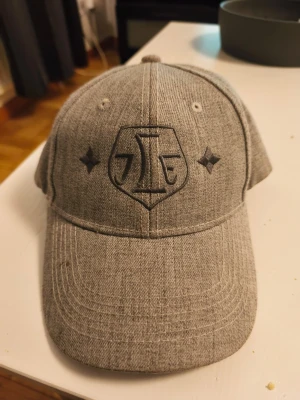 Grå keps med broderad logga - Snygg grå keps i mjukt tyg med böjd skärm och broderad logga framtill med Leksands IF och två stjärnor. Klassisk sexpanelsdesign och justerbar baktill. Perfekt för en avslappnad streetwear-look.