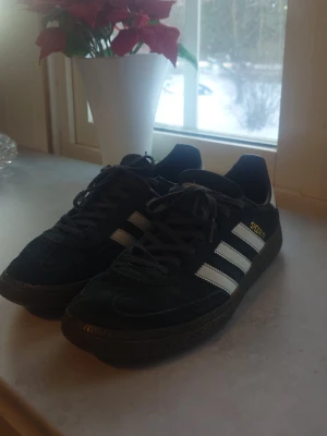 Adidas Spezial svarta sneakers - Säljer ett par svarta Adidas Spezial sneakers med klassiska vita ränder på sidorna och guldfärgad text. Skorna har låga skaft och ovandel i mocka med snörning. Perfekta för dig som gillar retrostil och streetwear.