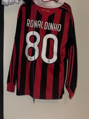 Retro AC Milan Ronaldinho tröja Adidas M - Säljer en klassisk AC Milan matchtröja med Ronaldinho 80 på ryggen. Tröjan är svart och röd med vertikala ränder, Adidas-logga och Bwin sponsortryck. Tillverkad i lätt polyester med Clima365-teknologi och broderat klubbmärke på bröstet. Perfekt för alla Milan-fans!