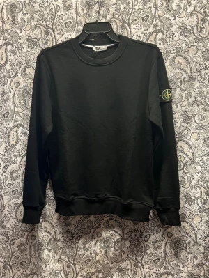 Svart sweatshirt från Stone Island - Säljer en svart sweatshirt från Stone Island med klassisk rund hals och ribbade muddar. Tröjan har den ikoniska Stone Island-loggan på vänster ärm. Perfekt för en clean och stilren look.