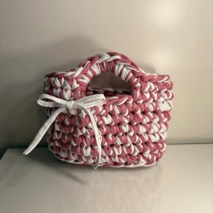 Virkad handväska i t-hirt garn i rosa/vit - •Virkad handväska som smyckas av en söt rosett  •Virkad i t-shirt garn •Färg: Rosa & Vit   •Storlek S Mått: ca 20 cm bred & ca 18 cm hög  •Obs: Storlekarna kan skilja lite,  beroende på garnets tjocklek.  •Virkad av mig: @virkningbynadja   •Kolla gärna in min Instagram, @virkningbynadja 