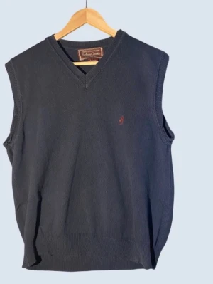 Mörkblå stickad ärmlös tröja från malboro classics medium - Klassisk mörkblå stickad väst från Brooks Brothers med v-ringning och diskret broderad logga i rött på bröstet. Västen är ärmlös och har ribbade kanter vid hals, ärmhål och nederkant. Perfekt att bära över skjorta för en stilren look.