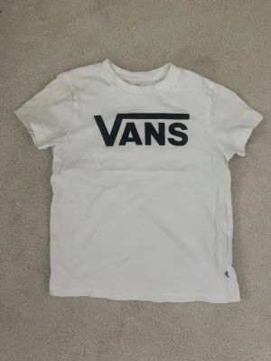 Vit Vans t-shirt med logga - Vit t-shirt från Vans med stor svart logga på bröstet. Klassisk rund halsringning och korta ärmar. Tillverkad i mjuk bomull för en skön känsla. Perfekt för en avslappnad och stilren look.