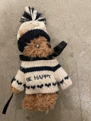 Nallebjörn med stickad tröja och mössa - Söt brun nallebjörn med fluffig päls, klädd i en beige och mörkblå stickad tröja med texten 'BE HAPPY' och matchande mössa med tofs. Tröjan har ett randigt mönster och nallen har ett svart band runt halsen. Perfekt som dekoration eller present.