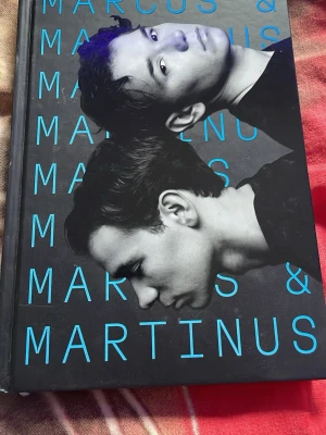 Marcus & Martinus bok med hård pärm - En snygg bok med svart och blått omslag, prydd med ett foto av Marcus & Martinus samt deras namn i stora blå bokstäver. Boken har hård pärm och insidan är blå. Perfekt för fans av popduon och dig som gillar biografier och skönlitteratur.
