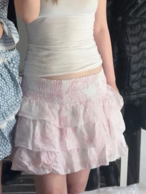 Volangkjol i rosa och vitt från Nelly - Supersöt kort volangkjol från Nelly i ljusrosa och vitt med elastisk smock i midjan. Kjolen har flera lager volanger och ett diskret blommigt mönster