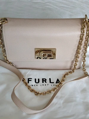 Ljusbeige axelväska från Furla - Snygg ljusbeige axelväska från Furla i läder med guldfärgad kedjerem och elegant metallspänne framtill. Klassisk rektangulär form och stilren design som passar till många outfits. Perfekt för dig som gillar stilrena accessoarer.