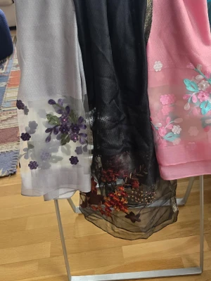 Blommiga sjalar i mesh och brodyr - Tre sjalar i mesh med broderade blommor i lila, rosa, turkos och rött. En är ljusgrå med lila blommor, en svart med röda detaljer och en rosa med pastellfärgade blommor och pärlor. Perfekta för att lyfta din outfit med färg och mönster.