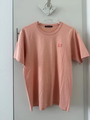 Rosa t-shirt från Acne Studios - Säljer en stilren rosa t-shirt från Acne Studios med klassisk passform och rund hals. T-shirten har en liten patch med Acne Studios-loggan på bröstet och är tillverkad i mjuk bomull. Perfekt basplagg för en enkel och clean look.