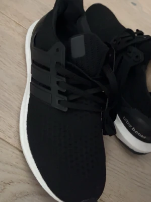 Svarta Adidas Ultra Boost sneakers - Snygga svarta Adidas Ultra Boost sneakers med vit sula och klassiska tre ränder på sidan. Ovandelen är i mesh för bra andningsförmåga och komfort. Perfekta för dig som vill ha både stil och funktion i vardagen.