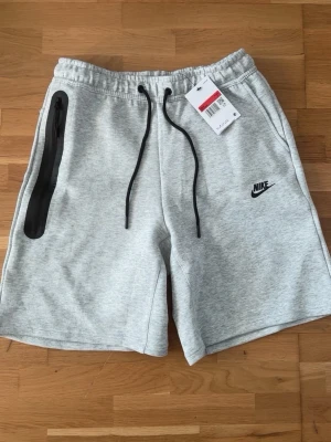 Grå Nike shorts med snörning - Säljer ett par ljusgrå shorts från Nike med svart logga på vänster ben. De har elastisk midja med svart snörning och en snygg dragkedjeficka på höger sida. Perfekta för sommarens aktiviteter och tillverkade i mjukt bomullsmaterial.