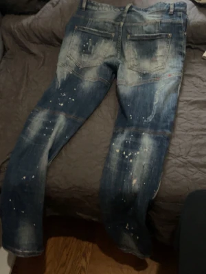 Dsquared2 blå distressed jeans - Säljer ett par blå jeans från Dsquared2 med coolt distressed och slitna detaljer samt färgstänk. Jeansen har raka ben, två bakfickor och är tillverkade i Italien. Perfekta för dig som gillar en edgy och trendig stil.