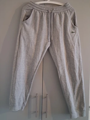 Grå mjukisbyxor&tröja från Lacoste set - Snygga grå mjukisbyxor från Lacoste med dragsko i midjan och sidofickor. Klassisk passform och broderad krokodil-logga på vänster sida. Tillverkade i mjukt bomullsmaterial för maximal komfort.