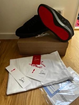 Svarta sneakers från Christian Louboutin - Snygga svarta sneakers från Christian Louboutin med den ikoniska röda sulan. Skorna har en klassisk låg siluett och är tillverkade i mocka med svarta skosnören. Kommer med originalkartong, dustbag och äkthetskort.