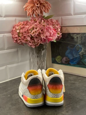 Nike Air Jordan 3 med färggradering - Snygga Air Jordan 3 sneakers i vitt läder med grått elefantmönster och färggradering i gult, orange och rött på hälen. Skorna har låga skaft, vita snören och detaljer i gult vid snörningen. Perfekta för dig som vill sticka ut med din stil.