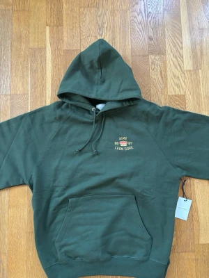 Grön hoodie från Aimé Leon Dore - Mörkgrön hoodie från Aimé Leon Dore med broderad logga och text på bröstet samt stor gul text och krona på ryggen. Klassisk känguruficka framtill, justerbar huva och ribbade muddar. Tillverkad i mjuk bomull för maximal komfort.