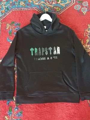 Svart Trapstar hoodie med tryck - Svart hoodie från Trapstar med stort tryck på bröstet i grönt och vitt där det står 'TRAPSTAR IT'S A SECRET'. Klassisk känguruficka framtill och huva. Perfekt för en streetwear-look.