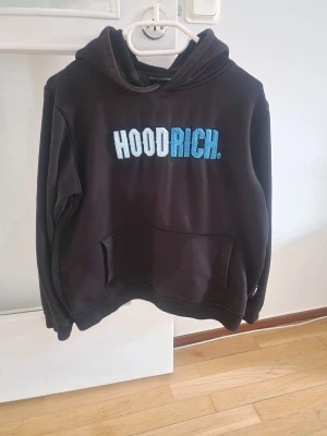Svart HOODRICH hoodie med brodyr - Säljer en svart hoodie från HOODRICH i storlek L (12-13Y). Hoodien har en stor broderad HOODRICH-logga i vitt och blått på bröstet, klassisk huva och en stor magficka framtill. Perfekt för dig som gillar streetwear och vill ha en skön och snygg hoodie.