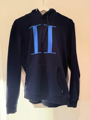 Mörkblå hoodie från Les Deux - Snygg mörkblå hoodie från Les Deux med stor blå romersk siffra II på bröstet. Klassisk känguruficka framtill, ribbade muddar och justerbar huva med snören. Perfekt för en avslappnad och trendig stil.